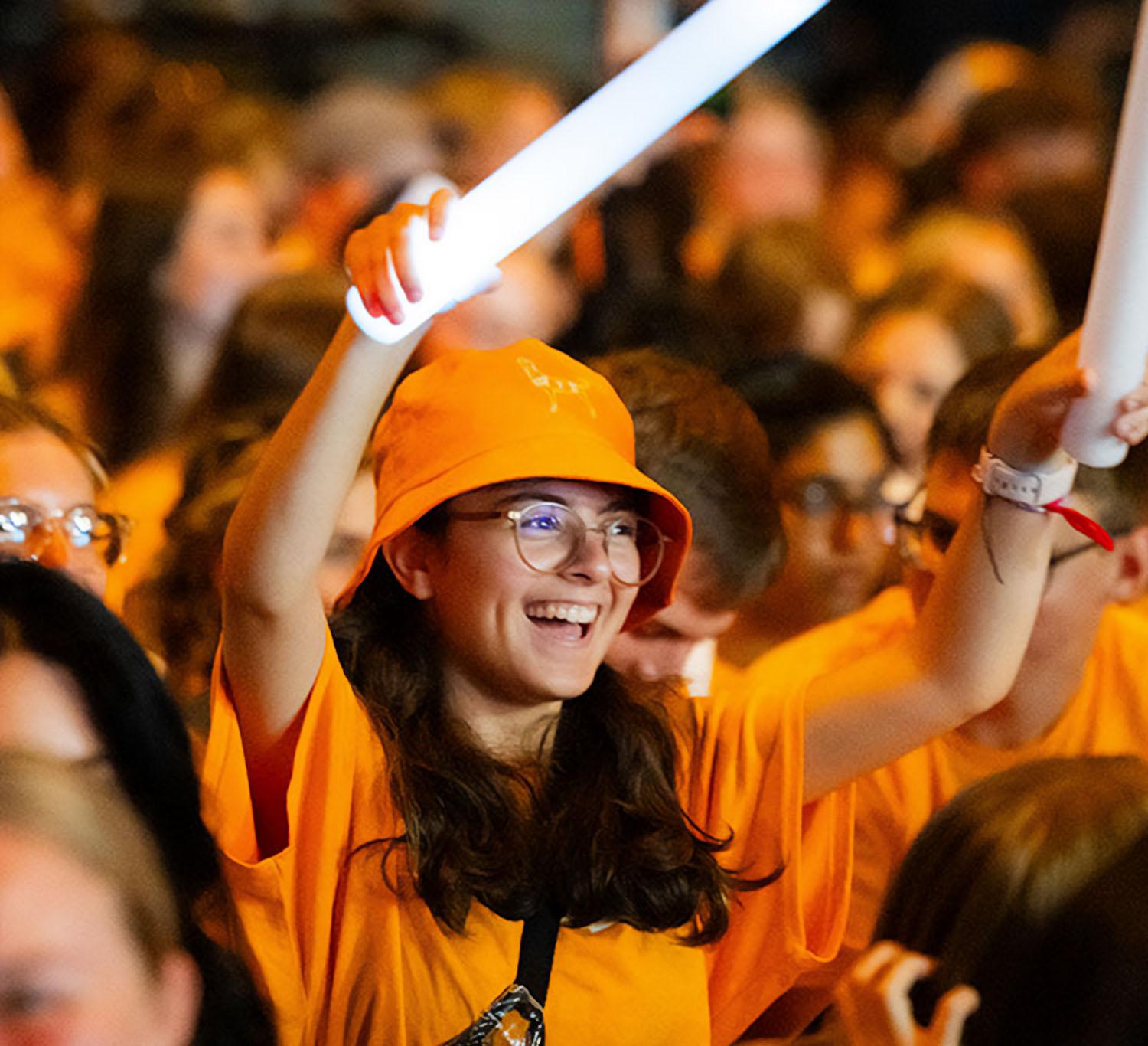 a smiling student holding large glow sticks in the middle of a sea of orange jams to a concert